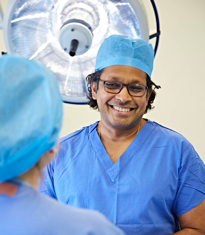 Man wearing a surgeon uniform, standing smiling to a colleague who has their back to frame